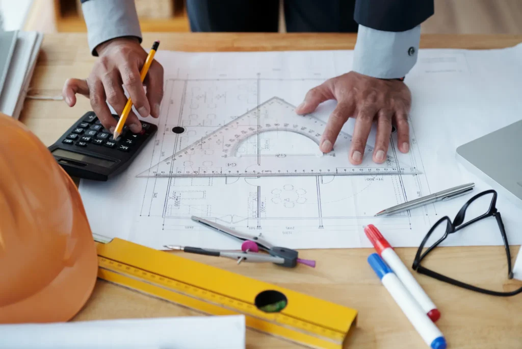 Close-up of an architectâ€™s hands measuring building plans with a triangular ruler while using a calculator, with drafting tools and a hard hat on the desk.