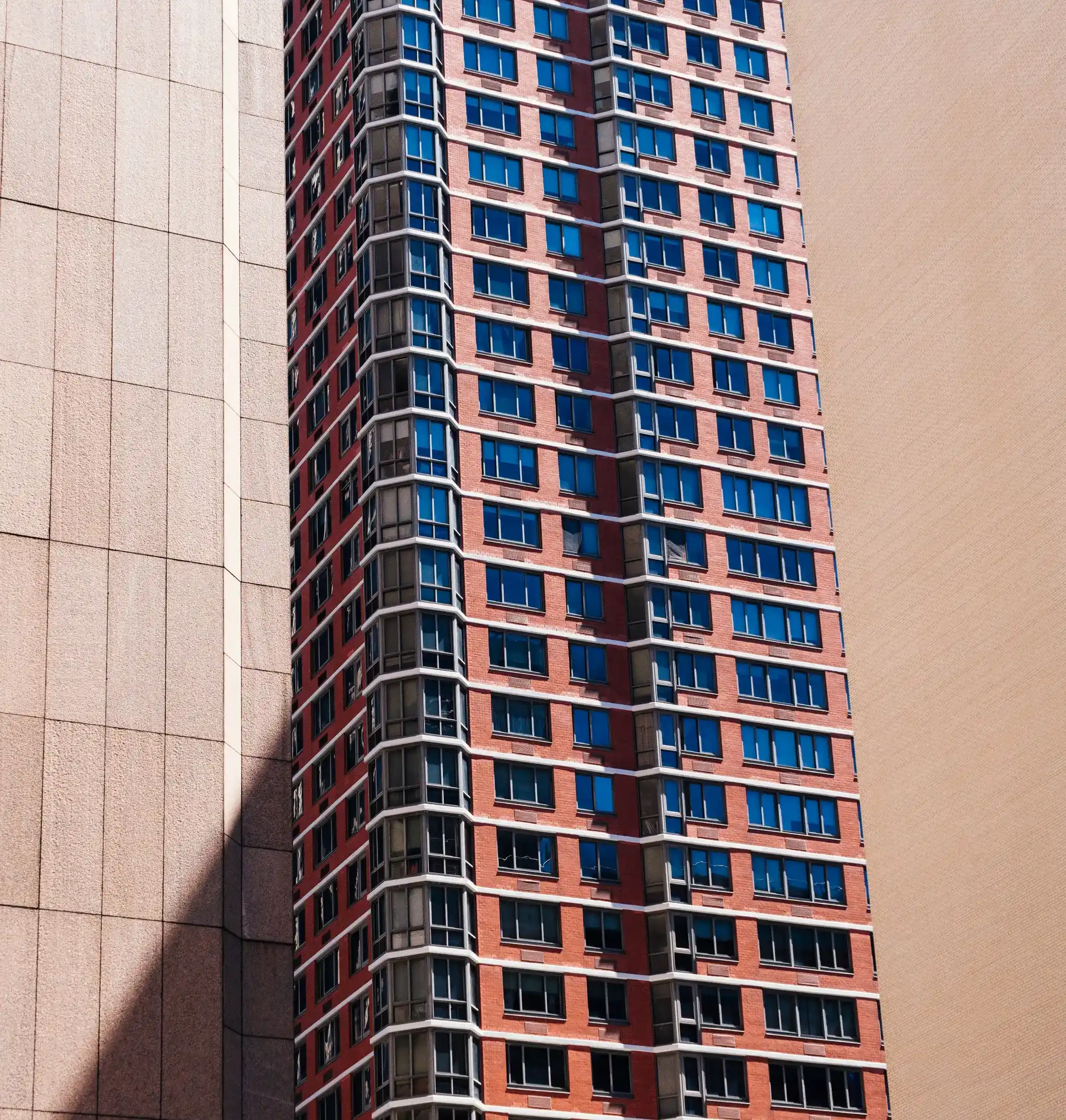 Tall red-brick high-rise with rows of blue-tinted windows, framed between two beige buildings.