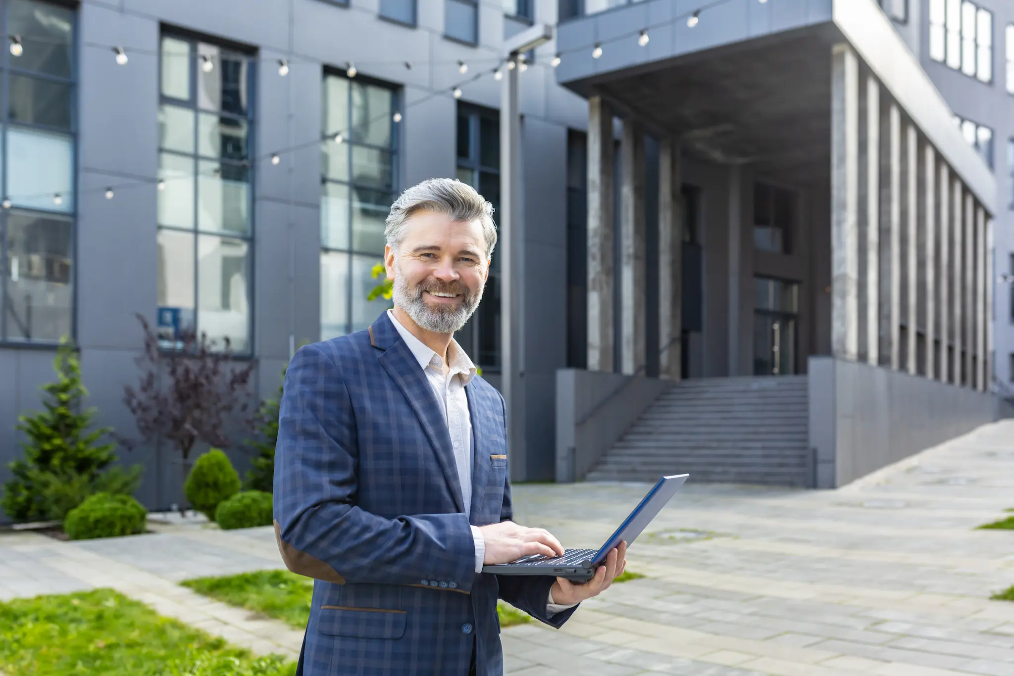 Smiling business professional standing outdoors with a laptop in front of a modern office building.