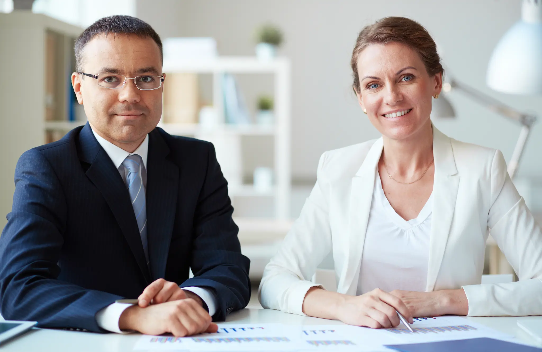 Two business professionals, a man in a dark suit and a woman in a white blazer, seated at a desk in a bright office, looking at the camera with documents spread in front of them.