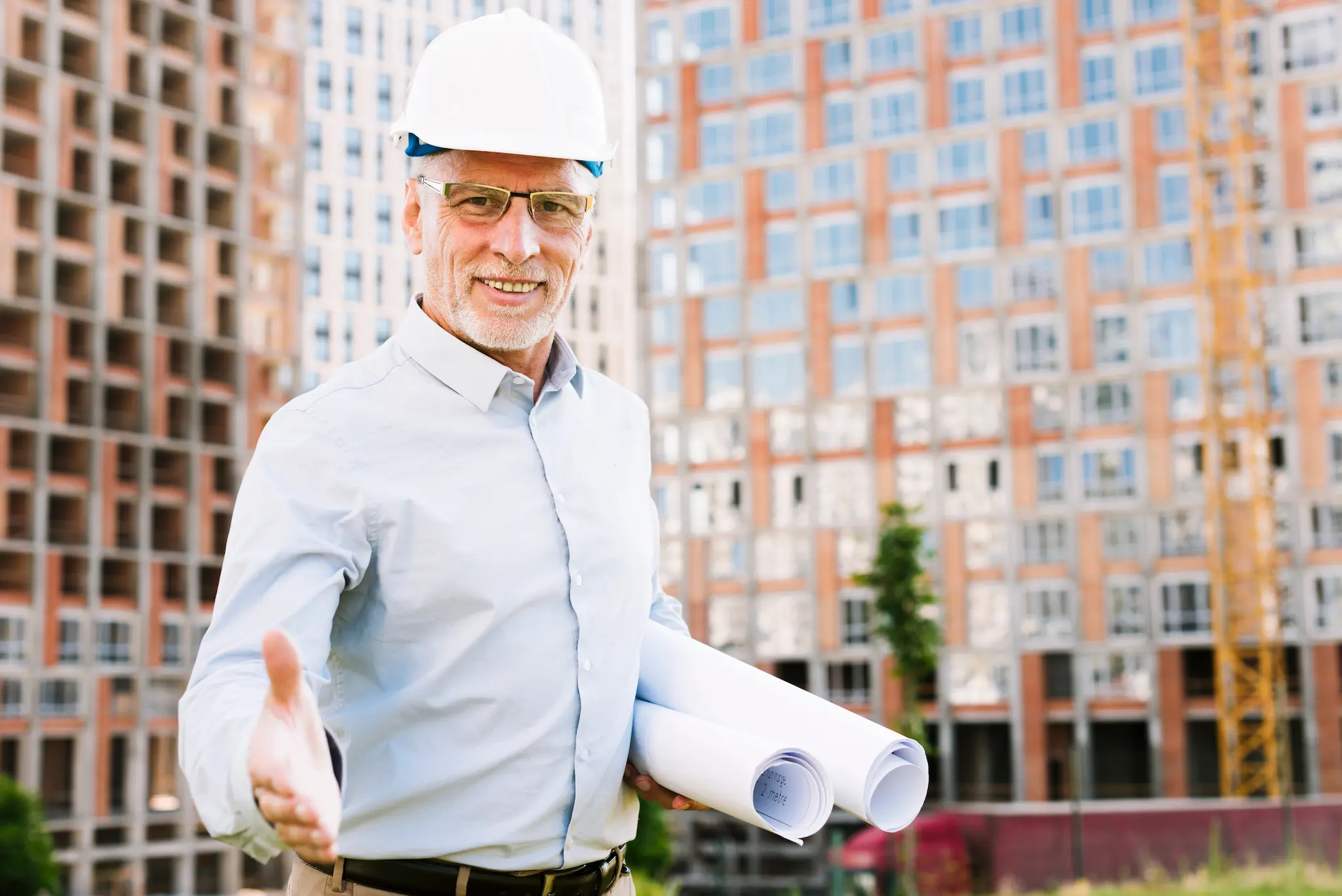 Smiling construction professional wearing a white hard hat and glasses, holding rolled blueprints and extending a hand in front of a residential building under construction.