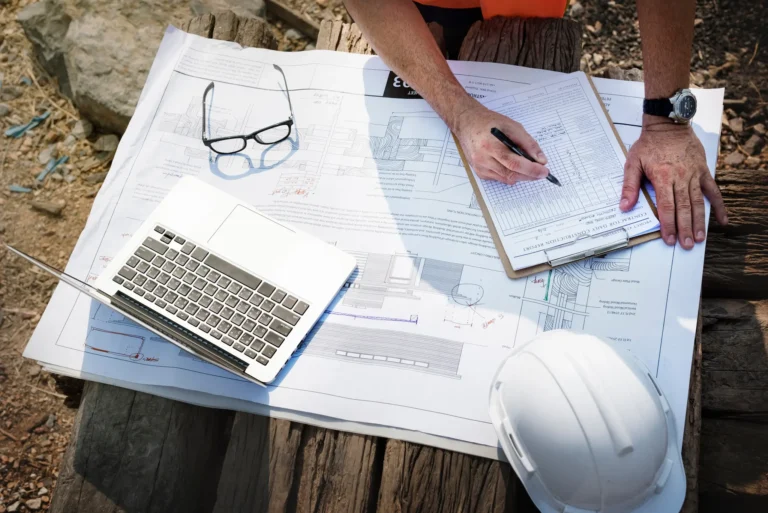 Person reviewing construction blueprints at an outdoor work site, with a laptop, clipboard, pen, and white hard hat placed on the plans.