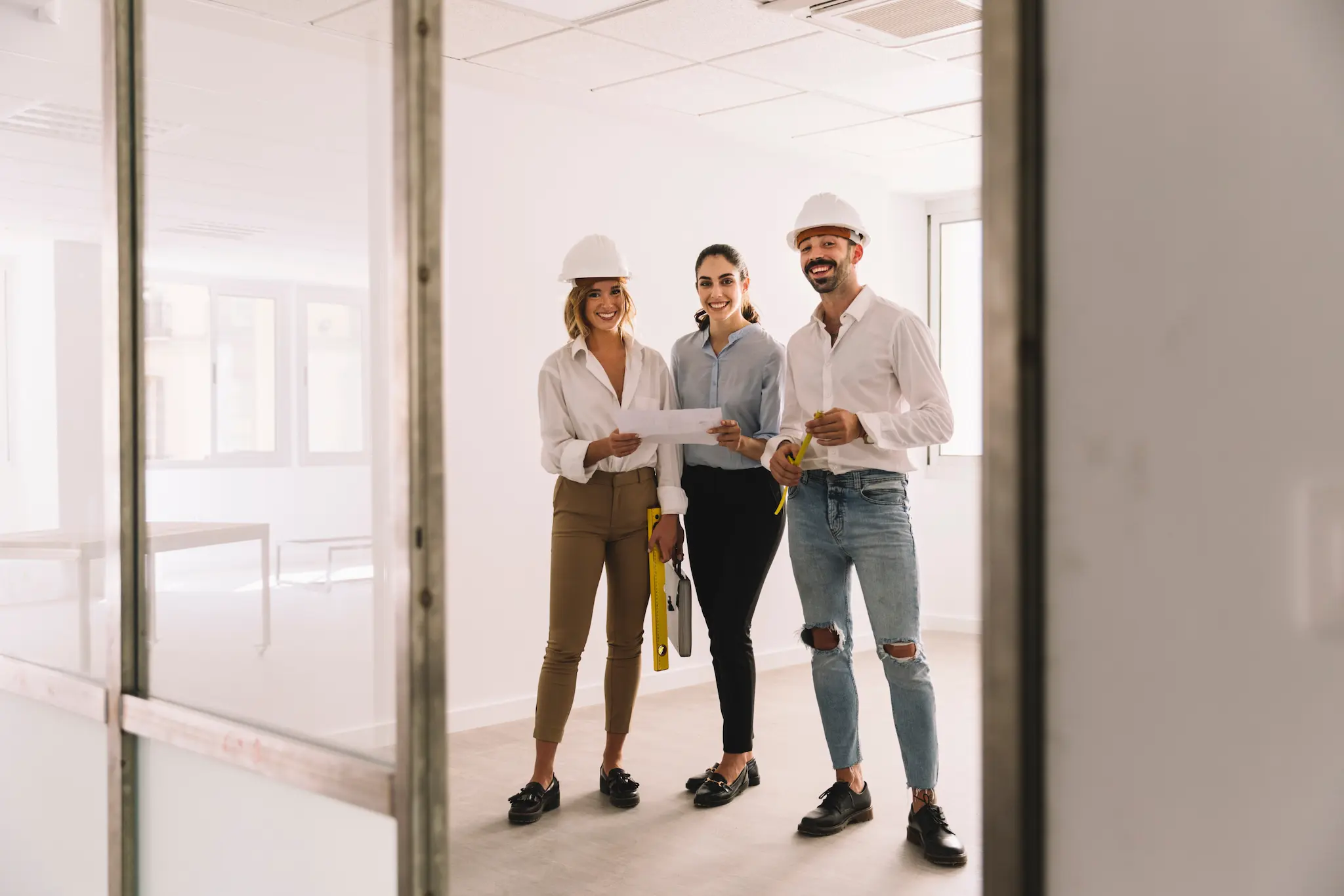Three professionals wearing hard hats stand inside a bright office space under renovation, holding plans and measuring tools while smiling at the camera.