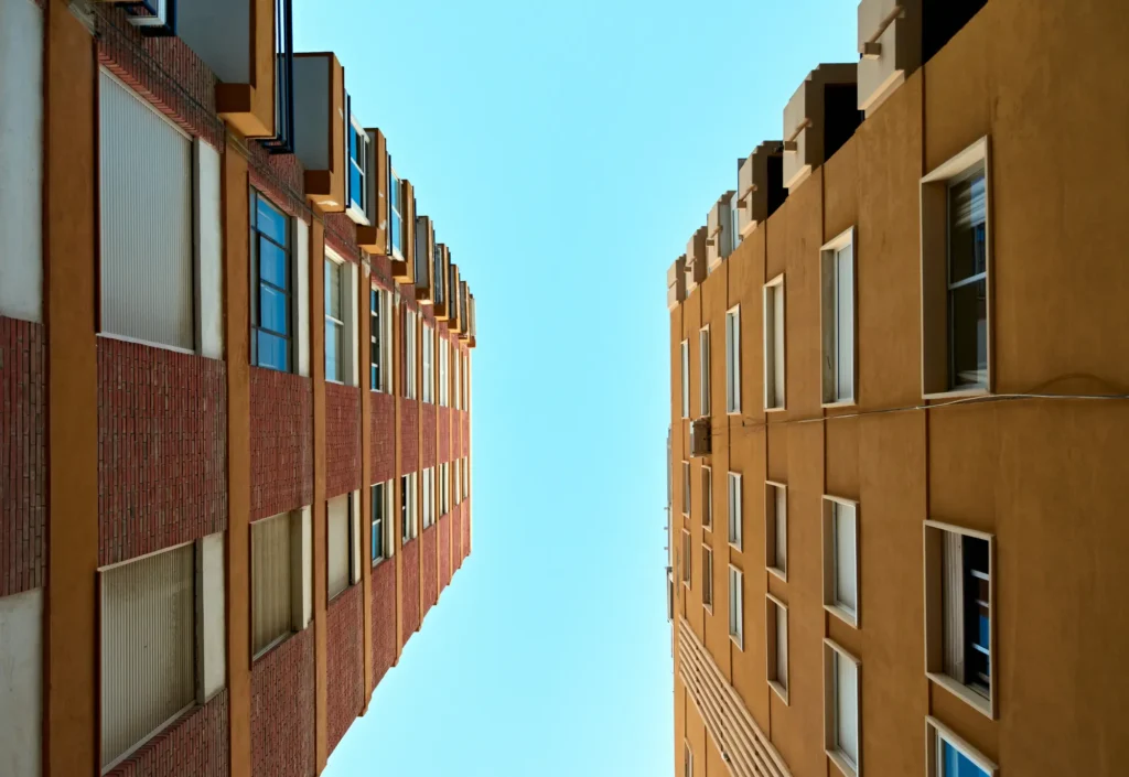 Upward view between two apartment buildings with rows of windows framing a bright blue sky.