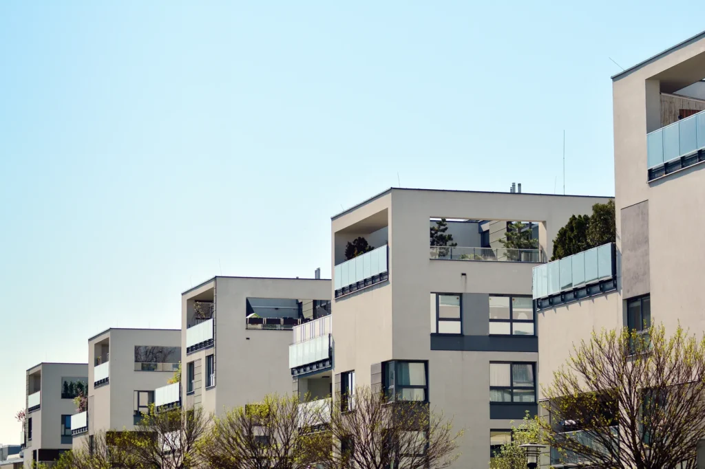 Row of modern apartment buildings with large windows and glass balconies under a clear blue sky, with trees in the foreground.
