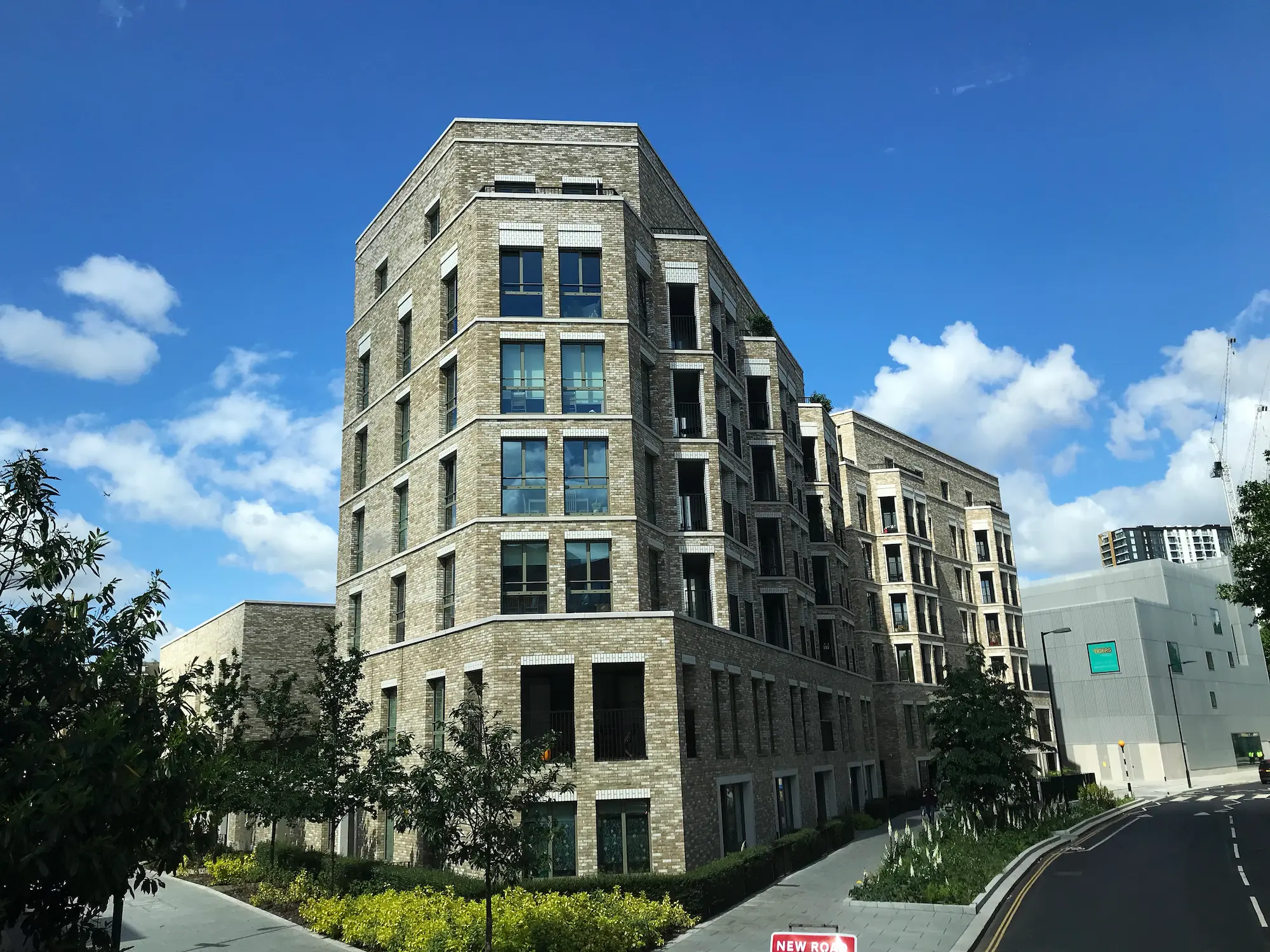 Modern mid-rise brick apartment building on a city street, with landscaped greenery and a clear blue sky.
