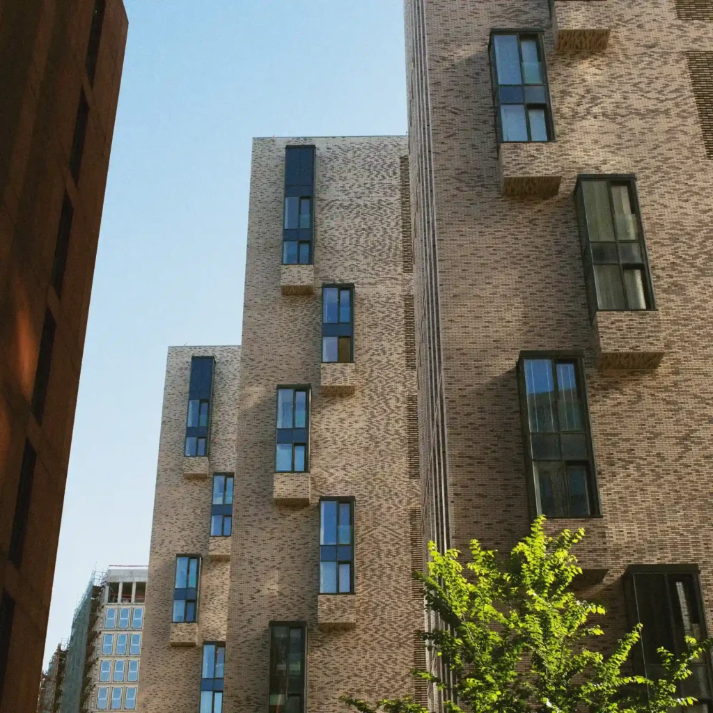 Tall modern brick apartment buildings with narrow windows rising against a clear blue sky, with a leafy green tree in the foreground.