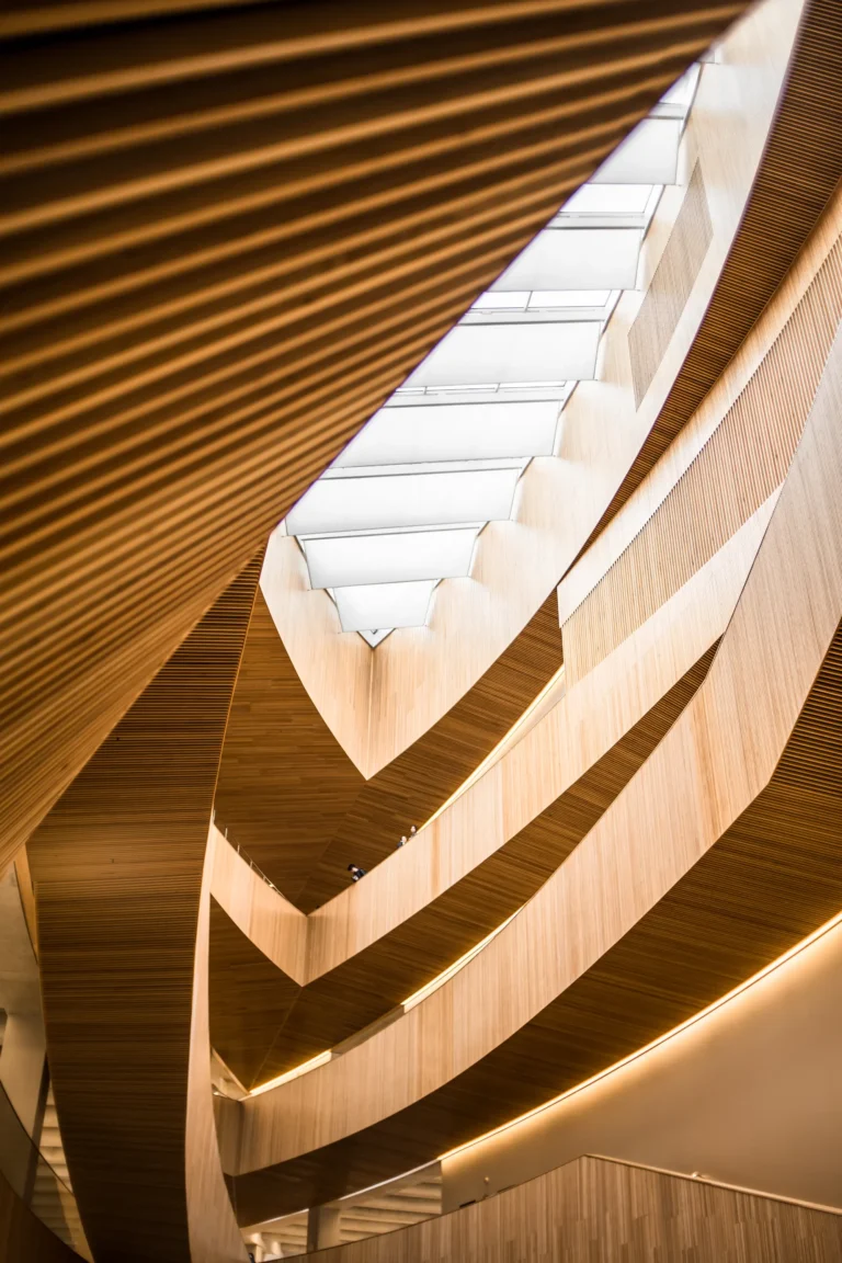 Interior view of a modern building with curved wooden walls, layered balconies, and a skylight ceiling creating sweeping architectural lines.