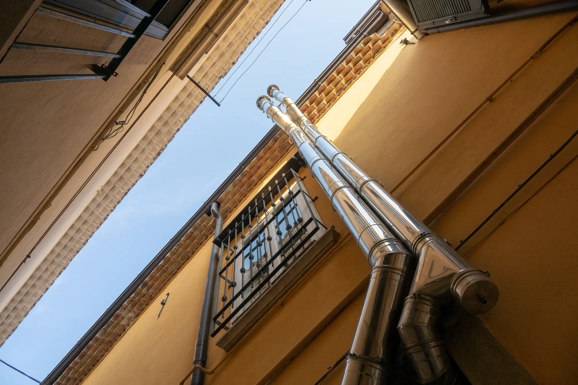 Upward view in a narrow alley showing shiny metal chimneys, yellow building walls, and a small balcony against a clear blue sky.