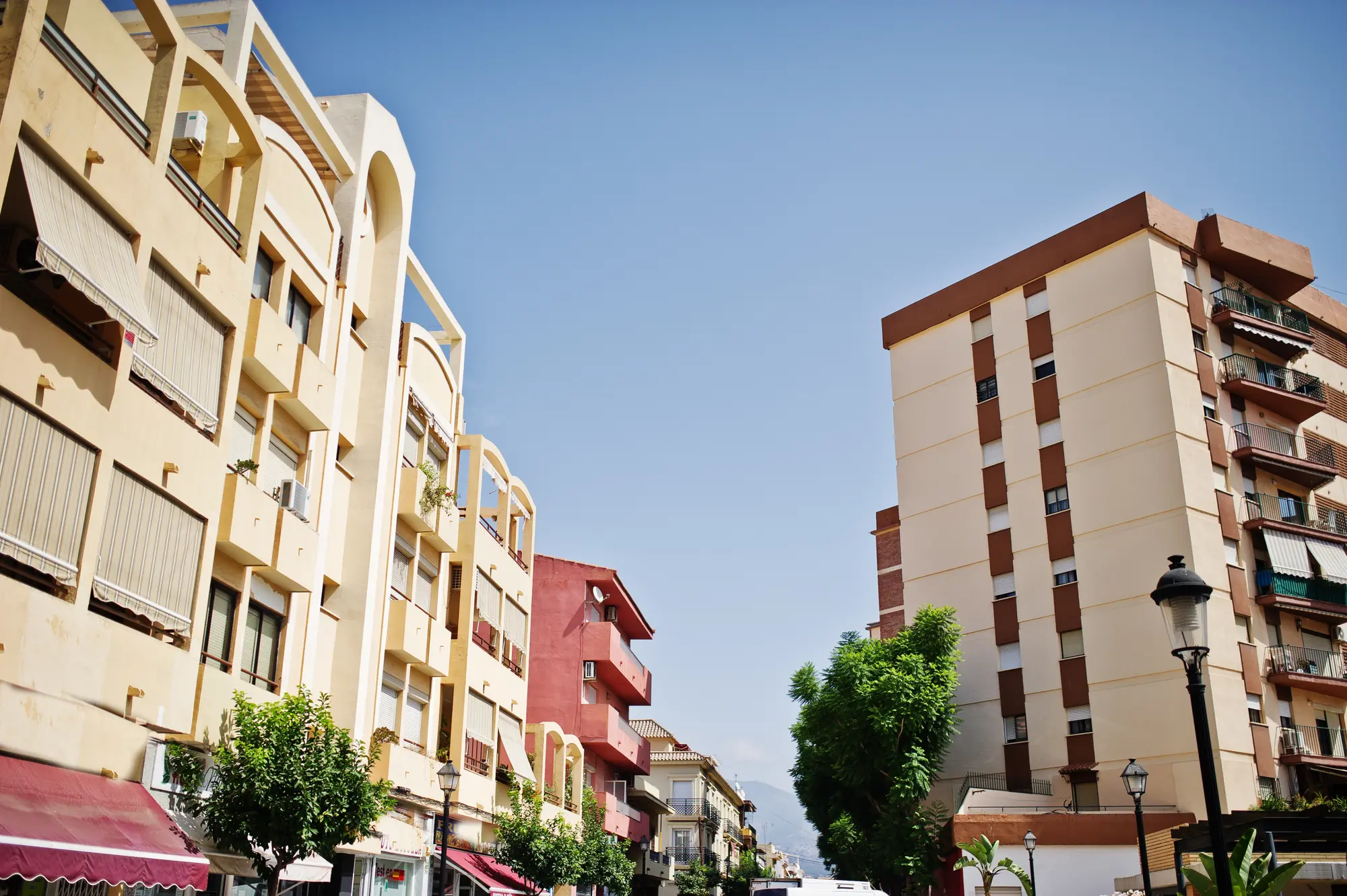 Sunny residential street lined with mid-rise apartment buildings, balconies, trees, and a clear blue sky overhead.