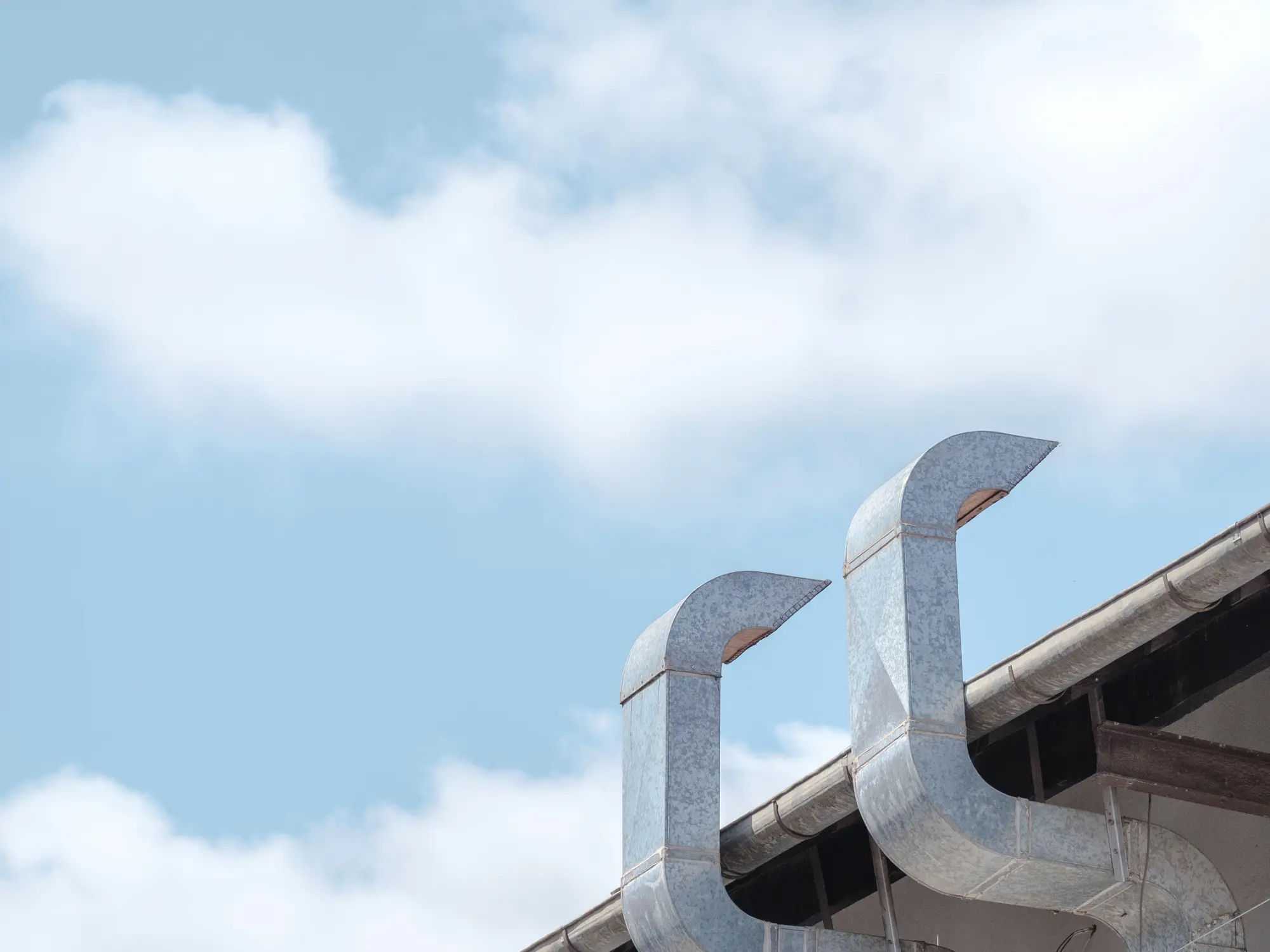 Two metal rooftop ventilation ducts extending upward against a bright blue sky with soft clouds.