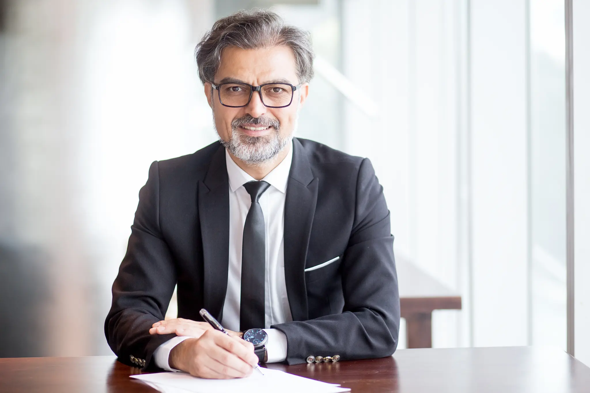 A professionally dressed man with gray hair and glasses, wearing a black suit and tie, sitting at a desk and smiling while holding a pen over a document in a bright office setting.